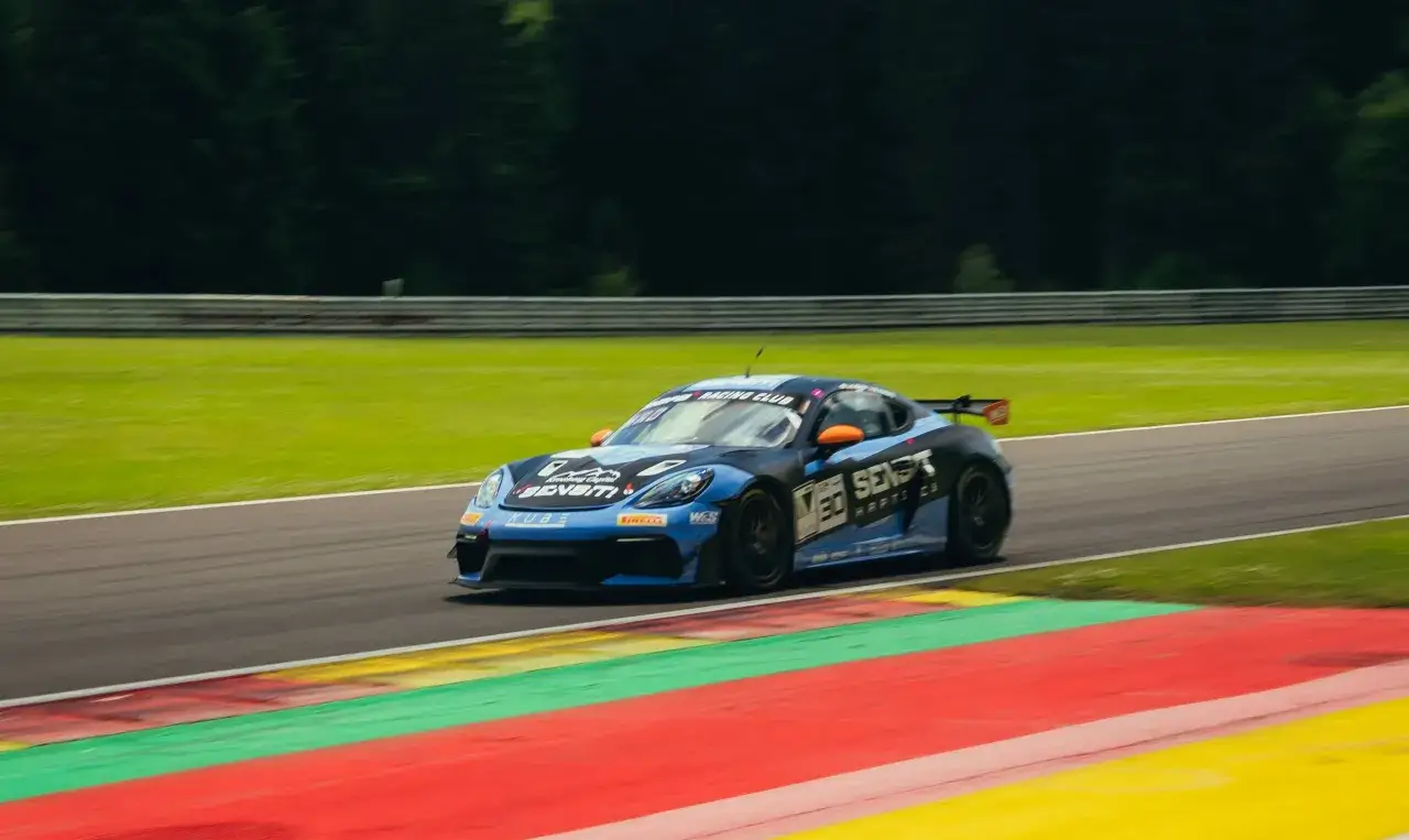 Niebiesko-czarny Porsche GT4 z pomarańczowymi akcentami pędzi po torze wyścigowym, zostawiając za sobą rozmyte pasy zieleni i czerwieni.
