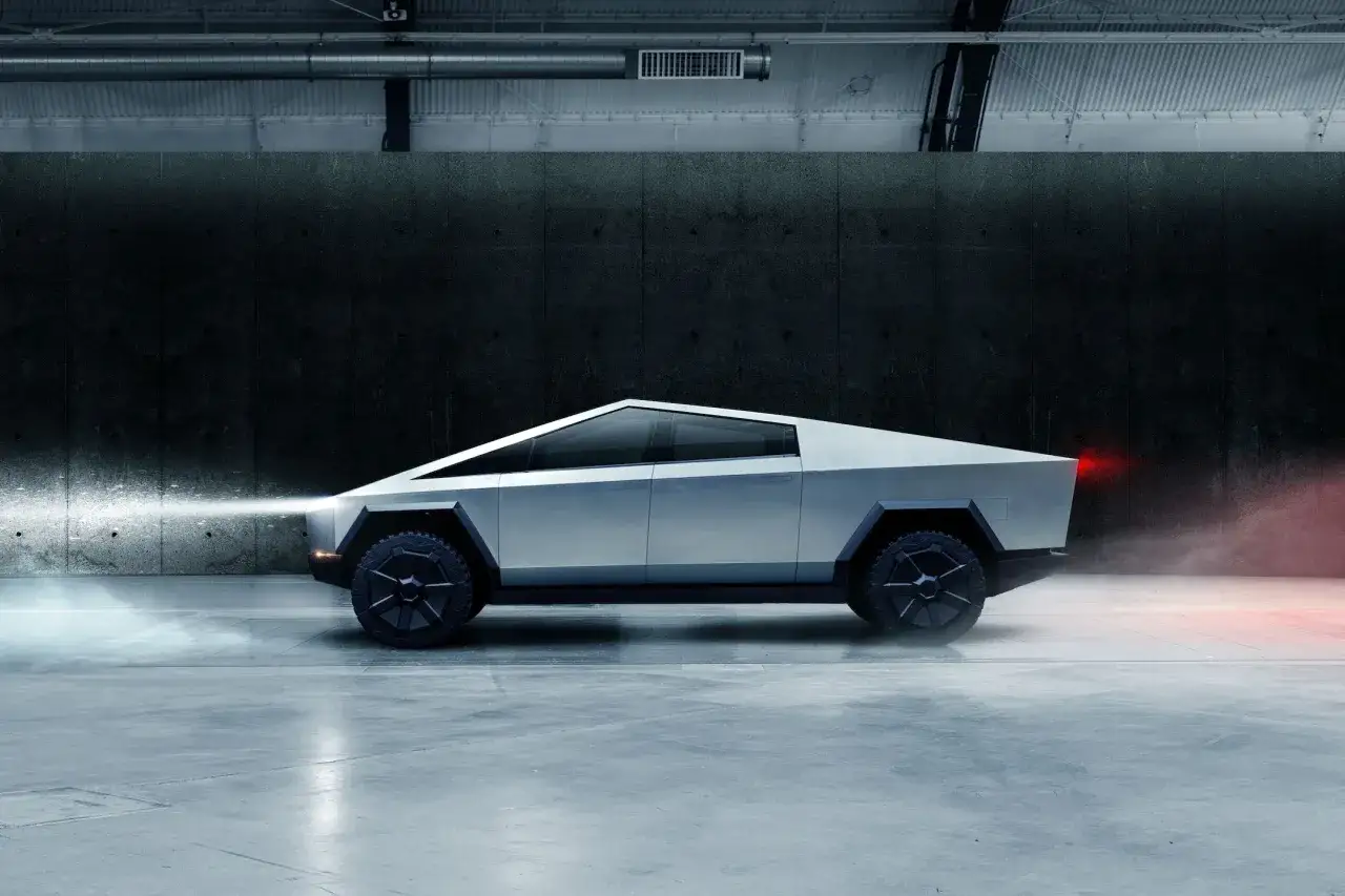 Futurystyczny Tesla Cybertruck z włączonymi światłami, unoszący się pył. Cena tego pojazdu jest wciąż przedmiotem spekulacji.