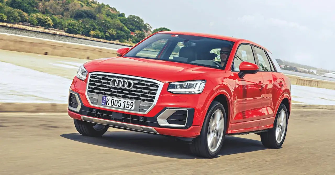Czerwony Audi Q1 w ruchu, z kierowcą w okularach przeciwsłonecznych za kierownicą, na tle krajobrazu z zielenią i budynkami.