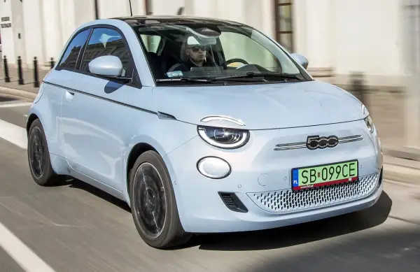 Jasnoniebieski fiat elektryczny 500e z czarnym dachem jedzie ulicą. Kierowca w okularach przeciwsłonecznych prowadzi auto.