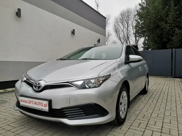 Srebrna Toyota Auris 1.6 benzyna zaparkowana na podjeździe z kostki brukowej, przed nowoczesnym budynkiem.