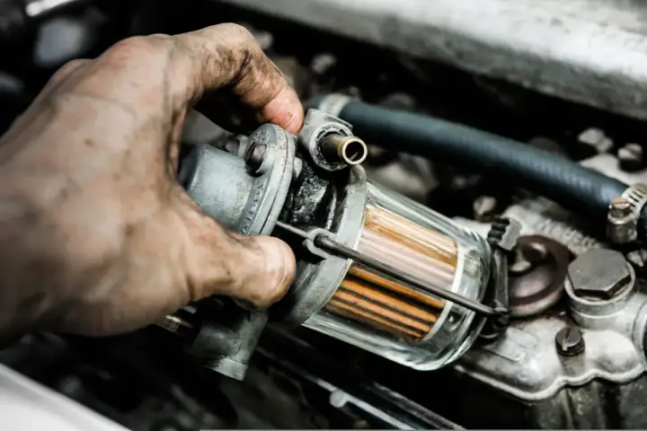 Brudna ręka mechanika wymienia jeden z filtrów w instalacji LPG. Widać przezroczysty korpus filtra z pomarańczowymi wkładami.